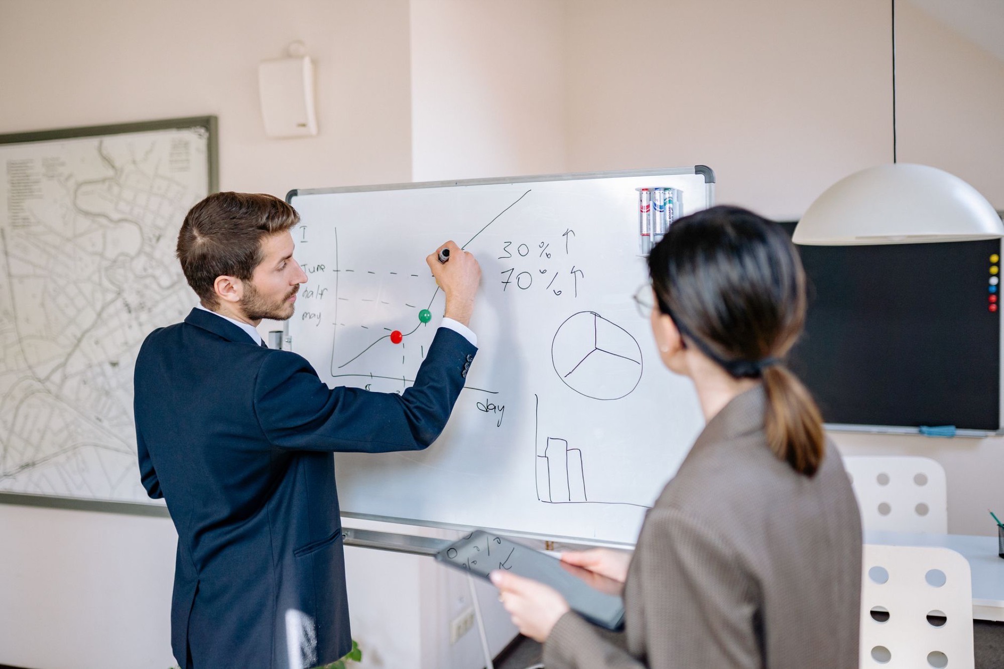 Two colleagues at a whiteboard in an office, one writing a line chart beside a hand-drawn pie chart and the figures 30% and 70%.